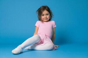 Adorable ballerina girl sitting on blue background and resting after ballet dance or choreography practice photo