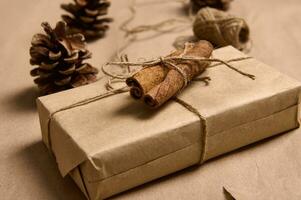 Beautiful simple present wrapped in craft paper decorated with tied cinnamon on the background of a rope and pine cones lying down on a brown cardboard. Christmas and New Year concept photo