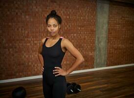 Confident portrait of a young African pretty woman, athlete with slim body, looks at camera posing with hands on waist at gym class with dumbbells and medicine ball lying on the floor photo