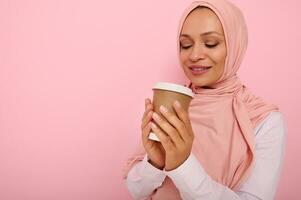 Beautiful Arabic muslim woman with covered head in pink hijab drinking hot drink, tea or coffee from disposable cardboard takeaway cup, standing three quarters against colored background, copy space photo