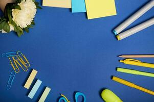 Flat lay of school office accessories in yellow shades, isolated on blue background, copy space. Aster flower and stationery supplies scattered in a circle. Back to school concept photo