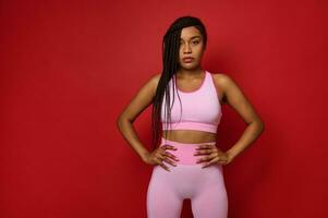 Self-confident beautiful young African American woman with stylish dreadlocks, wearing pink sportswear posing with arms on waist against a red colored background with copy space for advertisement photo