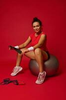Hispanic athletic woman holding elastic fitness band and sitting on fitness ball after exercising isolated on red background. Concept of slimness, healthy lifestyle and workout. photo