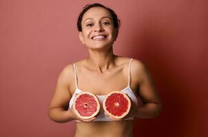 Beautiful woman in a white underwear holds grapefruits halves at chest level, smiling sweetly with a toothy smile posing on a coral background with space for text and advertising. Body and skin care photo