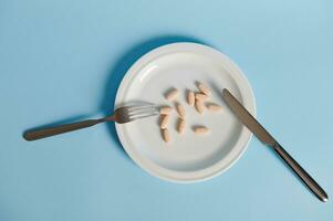 Flat lay composition with fork, knife and pills on a white plate. Isolated on blue background with copy space photo