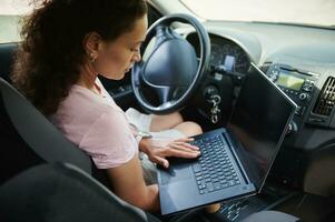 African-American woman sitting in the driver seat in personal car and working on laptop. photo
