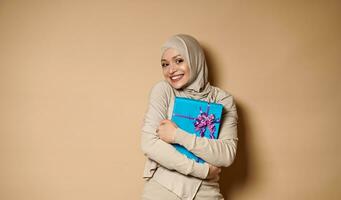 hermosa joven árabe musulmán mujer abrazos un regalo y sonrisas linda con dientes sonrisa a el cámara. beige fondo, Copiar espacio foto