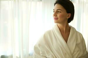 Serene attractive beautiful well-groomed young dark-haired woman with a stylish short haircut in a white terry bathrobe, smiling looking to the side against the background of the hotel room windows photo