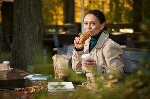 joven confidente hermosa negocio mujer disfruta su desayuno, come Fresco cuerno y bebidas café , resfriado lejos desde el ajetreo y bullicio de el ciudad, antes de distante trabajo en acogedor al aire libre café foto