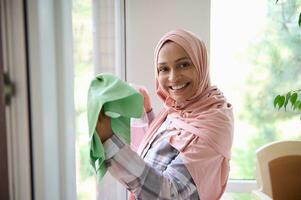 Arabic Muslim woman with head covered in pink hijab, smiles looking at camera, enjoying spring cleaning in the house on a sunny day, removing stains, washing windows in the veranda photo