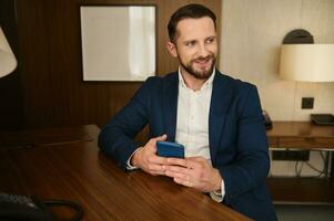 Smiling attractive businessman with mobile phone looking aside sitting at a table while resting in hotel room during business trip photo