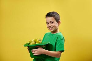 Side portrait of a smiling boy holding a leprechaun hat full of green apples. Saint Patrick's Day concept. Copy space. Yellow background. photo