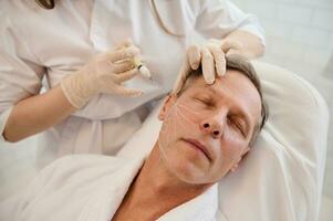 Beautician performs thread lifting according to the lines drawn on the mature handsome man's face. Rejuvenation, anti aging therapy concept for men in a modern spa clinic. Men's injection cosmetology photo