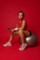 Beautiful athletic woman in red tight top and black sports shorts holding jump rope and smiling at camera while sitting on silver fit ball on red background with copy space photo