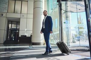 un longitud total hermoso empresario desarrollador empresario en un negocio traje con un maleta soportes a el Entrada de un moderno lujo edificio, caminando a el vestíbulo durante su negocio viaje foto