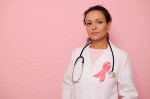 Confident portrait of a mixed race woman doctor in medical coat with pink ribbon, and stethoscope around neck, looking at camera, isolated on colored background, copy space. Breast Cancer Day concept photo