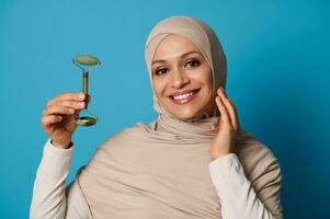 Smiling Muslim woman wearing a beige hijab holding a jade roller massager photo