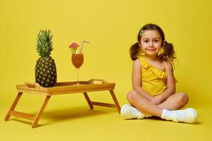 adorable pequeño niña sentado en amarillo antecedentes cerca un servicio bambú bandeja con un piña y cóctel. verano concepto foto