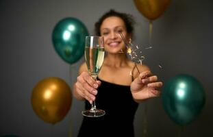 Soft focus on flute of champagne or sparkling wine and sparklers in the hands of happy woman having fun at anniversary, birthday party, Christmas New Year event. Gray background with air golden balls photo
