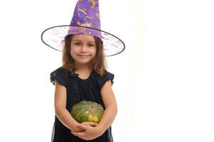 Víspera de Todos los Santos bruja concepto - retrato de pequeño caucásico niña vestido en elegante carnaval atuendo y mago sombrero, bruja niño sonrisas posando con calabaza en contra un blanco antecedentes con Copiar espacio foto