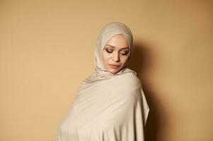 Portrait of young adult woman in muslim religious outfit and covered head looking down standing against a beige background with copy space photo