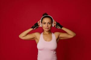 Charming Hispanic sportswoman puts on wireless headphones and confidently looks at camera, isolated on red background with advertising copy space photo