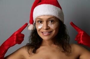 Attractive brunette in Santa costume points fingers on eye patches on her face, smiles looking at camera. Advertising for beauty salons for Christmas and New year giveaways. Spa, skin care concept photo