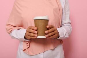 Close-up studio shot of female hands holding a craft takeaway cardboard cup with hot drink. Cropped view of woman in hijab with disposable paper mug, isolated on pink background with copy space photo