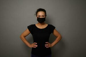 Isolated portrait on gray background of a woman in black medical protective mask looking at camera with hands on waist. Copy space for advertisement. Black Friday concept photo