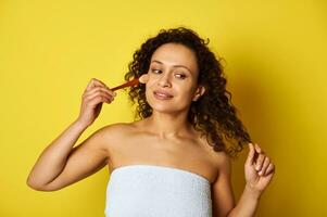 Charming half naked mulatto woman holding a makeup brush and applying blush on her face. Isolated beauty concepts on yellow background with copy space photo