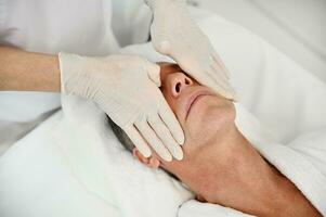 Close-up of female doctor's hands in white surgical gloves performing facial massage to a handsome mature man, patient in wellness spa clinic. Male beauty treatment and professional skin care concept photo