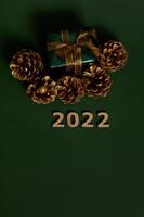 Top view of Christmas present in glitter wrapping paper with golden bow and pines cones colored in gold, numerals in wood symbolizing the year 2022 laid out on dark green background with copy space photo