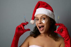 Sexy pretty woman in Santa hat, wears under eyes patches, rejoices looking at camera, holds syringes with beauty injection against gray background. Copy space for Christmas ad. Anti-aging cosmetology photo