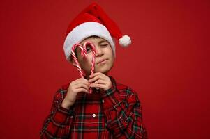 Cute boy in Santa hat and plaid shirt holds lollipops sugary striped candy canes in the heart shape and looks through it at the camera posing against red background with copy space for Christmas ad photo