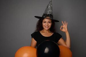 Beautiful smiling Hispanic woman in wizard hat, dressed in witch carnival costume for Halloween party, shows OK sign, poses against gray background with black balloons, copy space photo