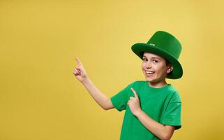 Funny little boy in Irish leprechaun hat with his finger pointing and looking at camera standing on yellow background with copy space. Saint Patrick Day photo