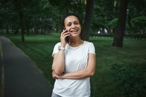 Portrait of a beautiful young woman talking on mobile phone while walking along the alley of a city park on a summer day photo