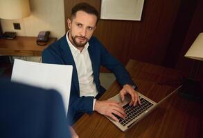 hermoso confidente negocio hombre, joven empresario trabajando en ordenador portátil sentado a un mesa en hotel habitación y mirando a su borroso jefe en el primer plano foto