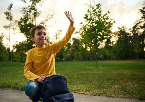 A friendly handsome friendly charming schoolboy 9 years old, sits on his knees on a path in the park, next to his backpack waves his hand, looks around and smiles sweetly with a toothy smile. photo