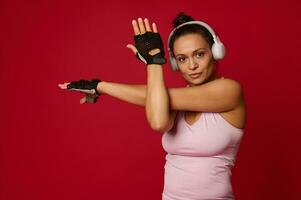 Confident active middle-aged active woman in sportswear and headphones stretching arms, looking at camera against red background with copy ad space photo