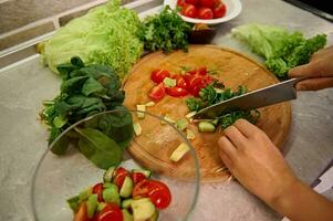 Female chef chopping raw vegetables and arugula leaves on a wooden kitchen board, preparing raw vegan salad at home kitchen photo