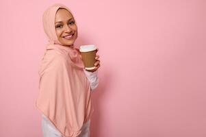 Arábica musulmán hermosa mujer con cubierto cabeza en hijab soportes Tres cuarteles en contra rosado antecedentes con para llevar taza desde desechable cartulina de caliente beber, sonrisas mirando a cámara. Copiar espacio foto