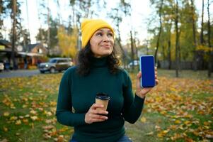 Pensive young woman stands in autumn forest park with cardboard cup of coffee and looks thoughtful up showing mobile phone with empty blank blue chroma key screen with copy space to insert ad. Mockup photo