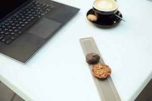Focus on delicious pastries on the white table next to your laptop and a cup of delicious aromatic cappuccino coffee. Copy space. photo