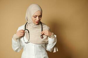 Portrait of a muslim woman doctor wearing hijab and putting a phonendoscope around her neck. Beige background with copy space photo