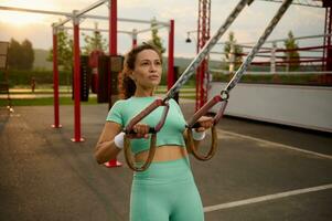 de cerca retrato de un bonito determinado africano americano mujer formación brazos con estacionario aptitud correas al aire libre, haciendo Lagartijas, formación Superior cuerpo, pecho, espalda, pectoral músculos, triceps foto