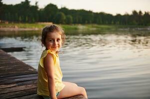 sonriente y contento niño niña sentado en un muelle a un lago a el verano noche. foto