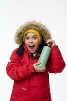 Vertical portrait of a cheerful school-age Caucasian boy winter traveler adventurer holding thermos flask and happily looking at camera, isolated on white background with copy space for advertisement photo