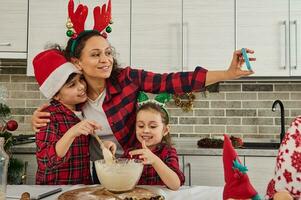 Adorable Caucasian children kneading dough, helping their mother in cooking Christmas bread and cookies. Delighted mom recording video and making selfie, hugging her son and daughter. Happy family photo