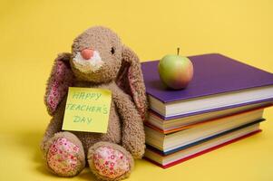 A plush toy rabbit with lettering Happy Teacher's Day on yellow note paper, leaned on a stoke of colorful books, yellow background with space for text photo